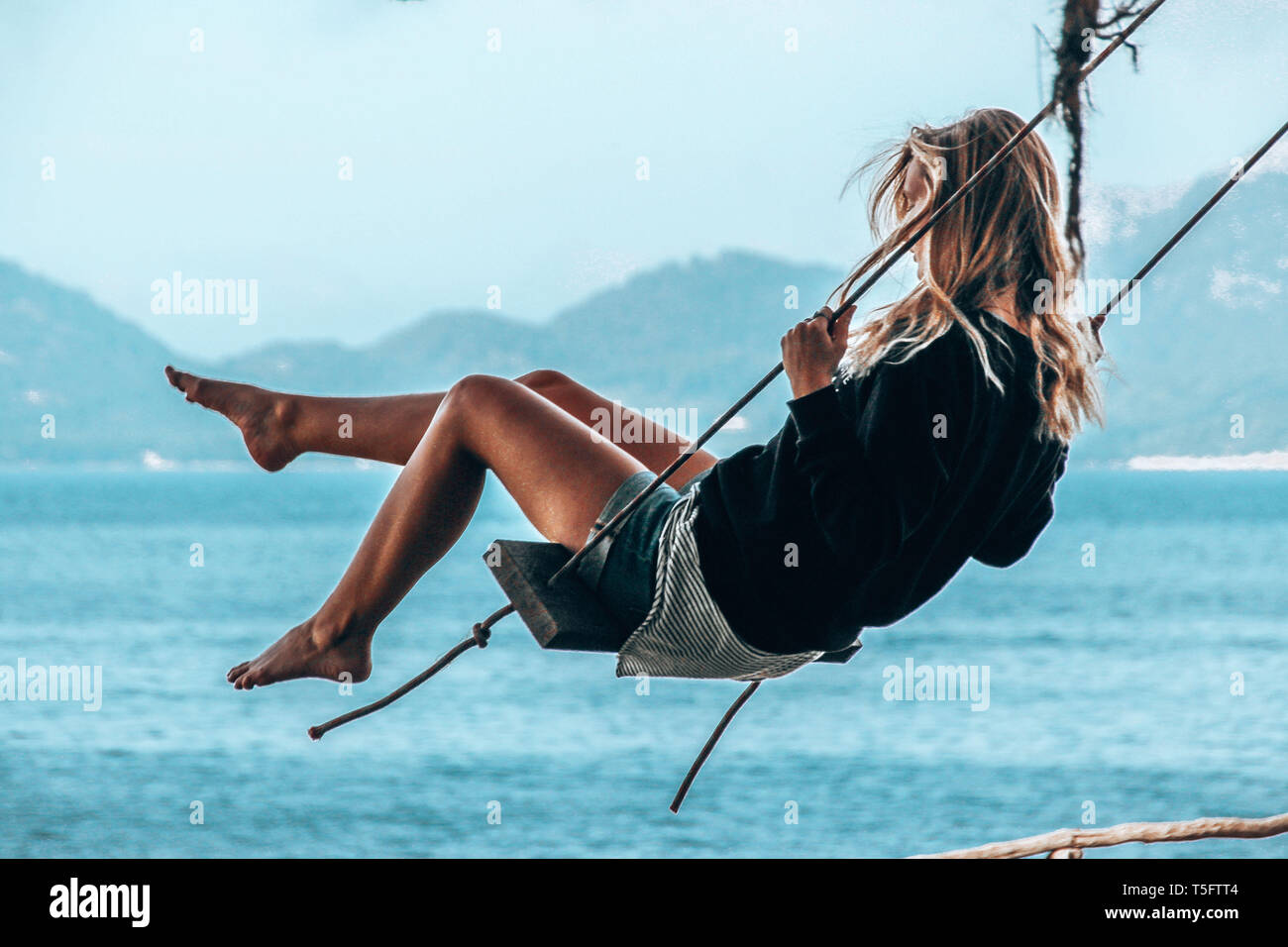 Ragazza donna seduta su una oscillazione dall'oceano con gambe lunghe e capelli biondi Foto Stock