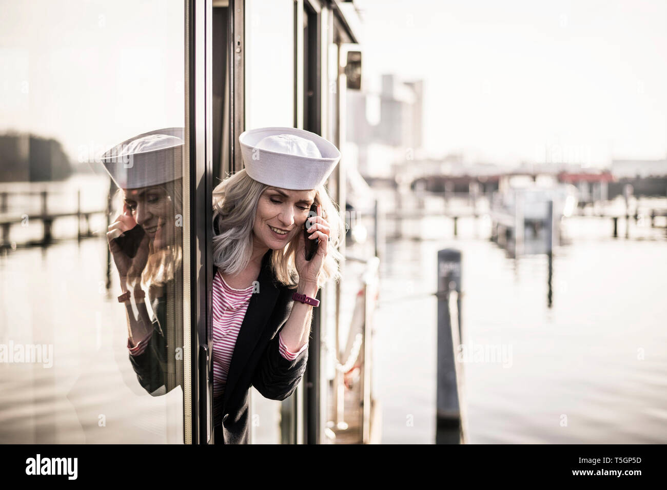 Imprenditrice indossando sailor hat, lavorando su una casa galleggiante, utilizza lo smartphone Foto Stock