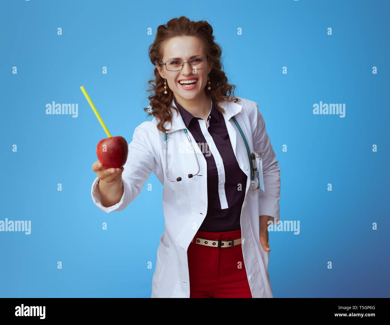 Sorridente medico moderno donna in bue t-shirt, pantaloni rosso e bianco manto medico dando un apple con cocktail verde tubo su sfondo blu. Foto Stock