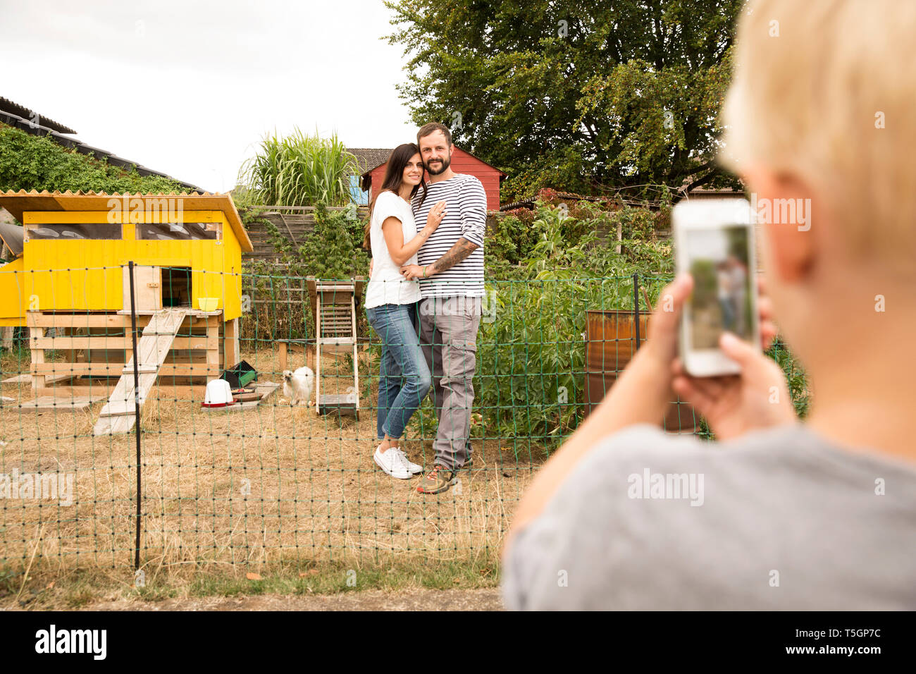 Ragazzo tenendo cellulare foto dei genitori permanente al chickenhouse in giardino Foto Stock