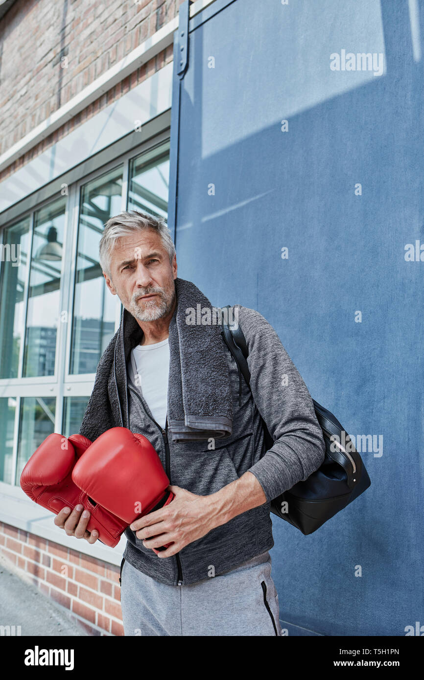 Ritratto di uomo maturo con asciugamano, borsa sportiva e rosso guantoni da pugilato in piedi nella parte anteriore della palestra Foto Stock