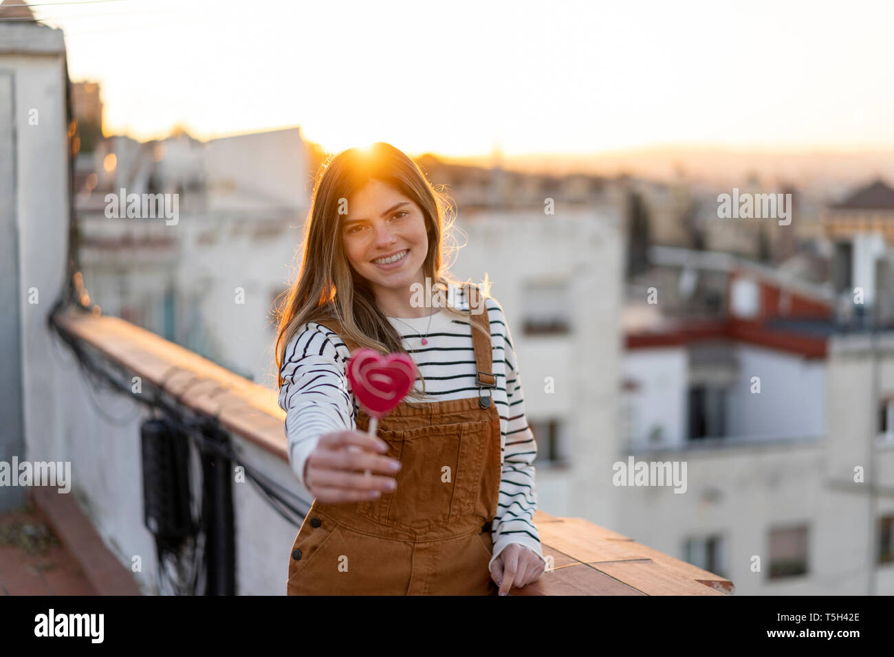 Ritratto di sorridente giovane donna gifting red lollipop sulla terrazza sul tetto al tramonto Foto Stock