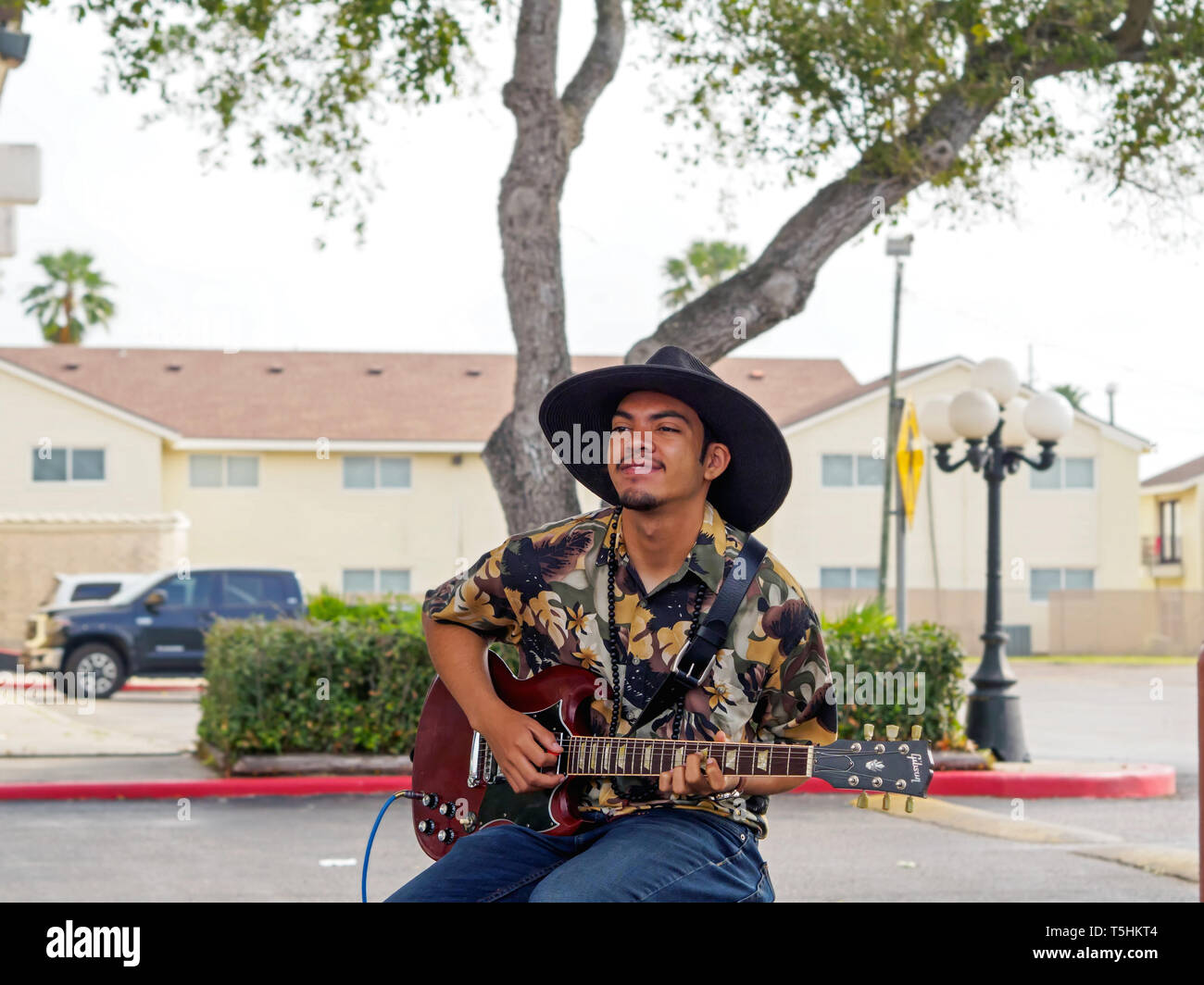 Un bel giovane con carnagione scura in un nero, largo-colmato hat riproduce una chitarra elettrica al Corpus Christi Southside Mercato degli Agricoltori. Foto Stock