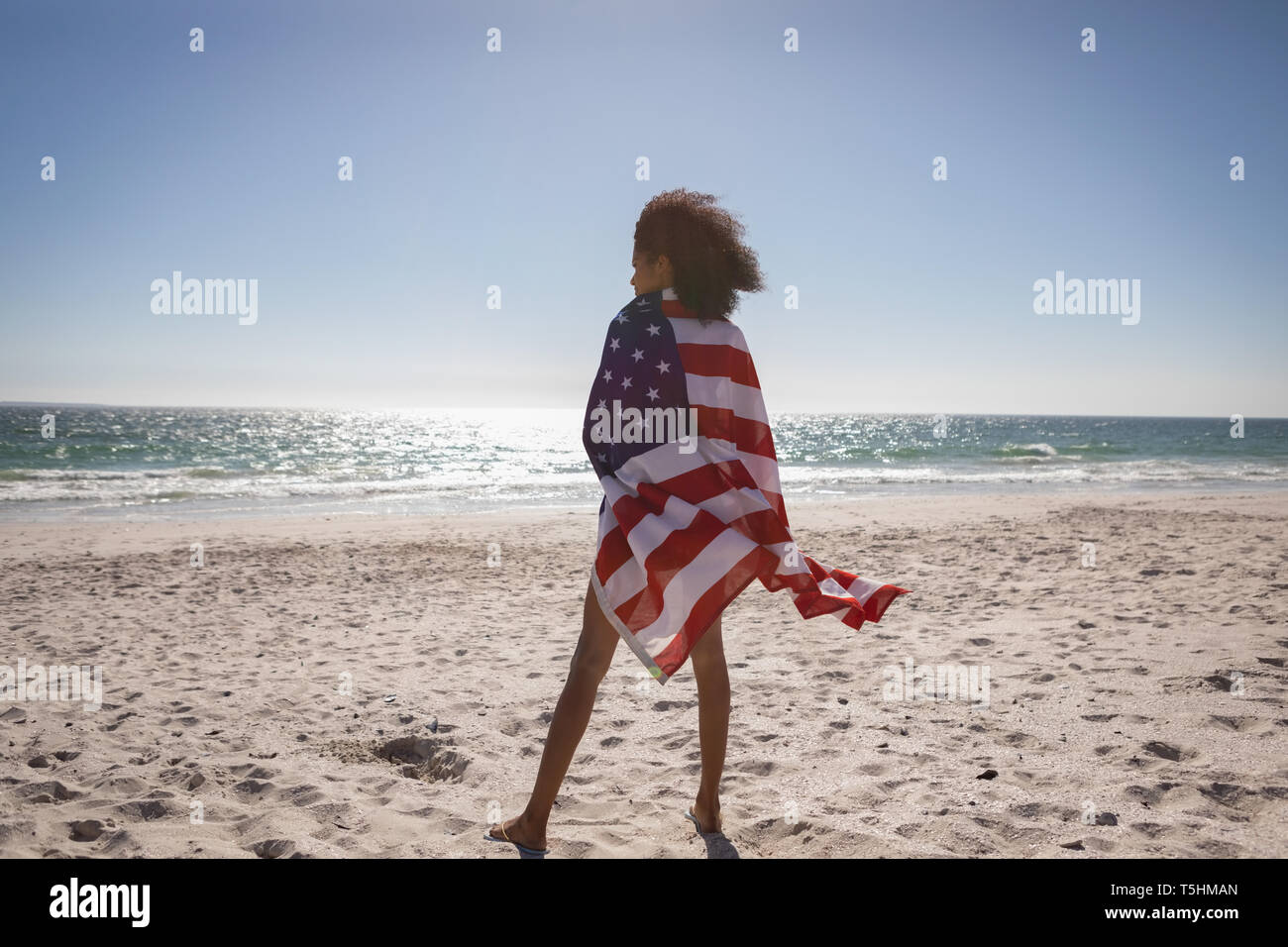 Bella donna avvolta bandiera americana mentre si sta in piedi in spiaggia in una giornata di sole Foto Stock