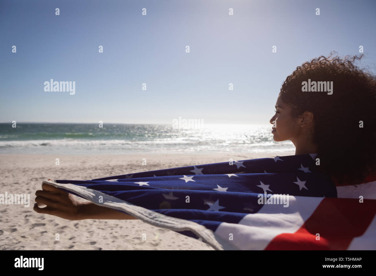 Bella donna holding bandiera americana mentre si sta in piedi in spiaggia in una giornata di sole Foto Stock