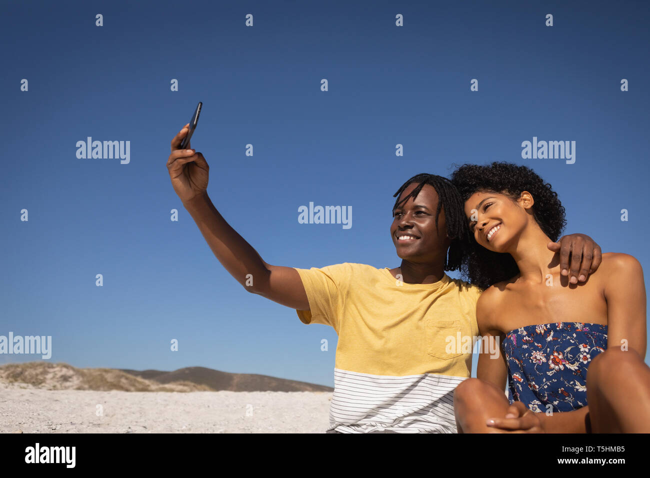Giovane tenendo selfie mentre è seduto alla spiaggia in una giornata di sole Foto Stock