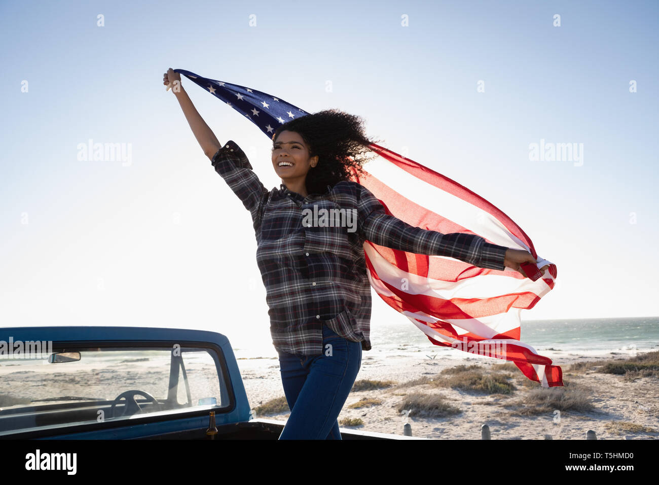 Bella donna holding bandiera americana mentre in piedi su auto in spiaggia Foto Stock