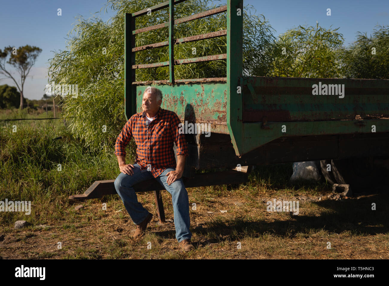 Senior uomo in attesa mentre è seduto nella parte posteriore del carrello in una fattoria in una giornata di sole Foto Stock