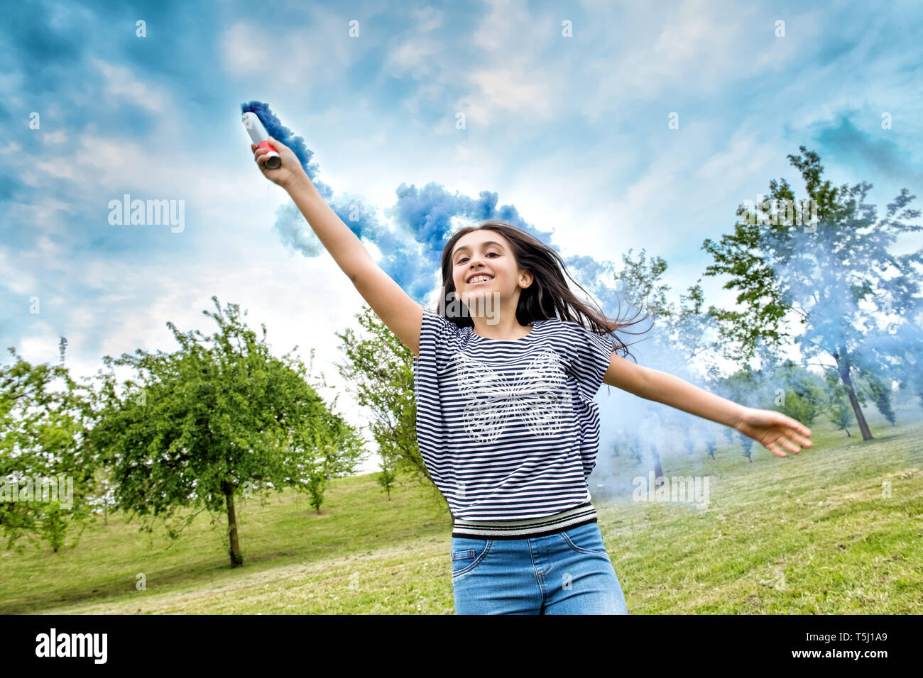 Felice di ridere ragazza giovane trailing una svasatura di fumo in una nuvola di coloratissimi fumo blu come lei corre verso la telecamera tenendola aloft in un parco a molla Foto Stock