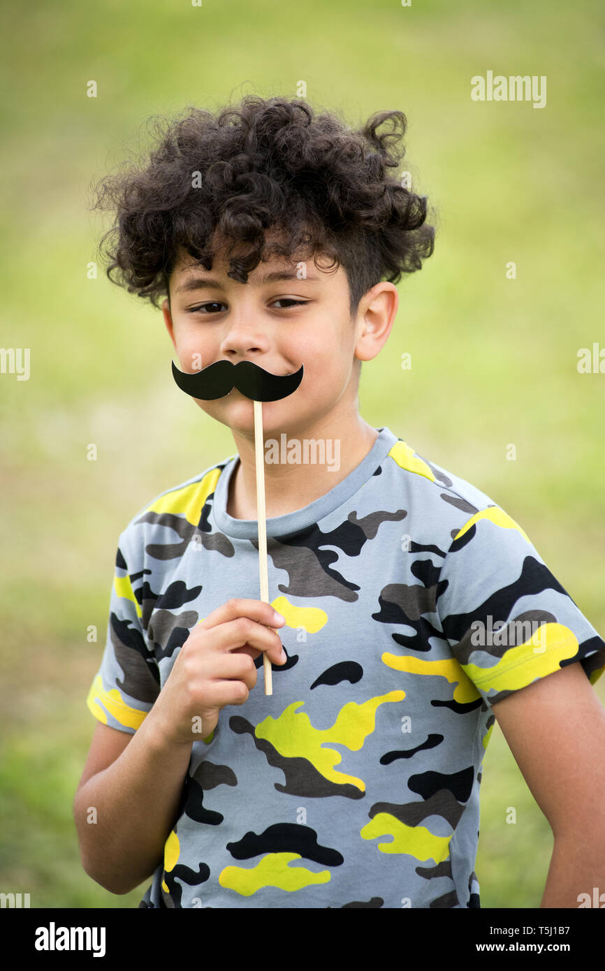 Giocoso ragazzino tenendo in mano un partito baffi accessorio con occhi sorridenti all'aperto contro sfocata erba verde Foto Stock