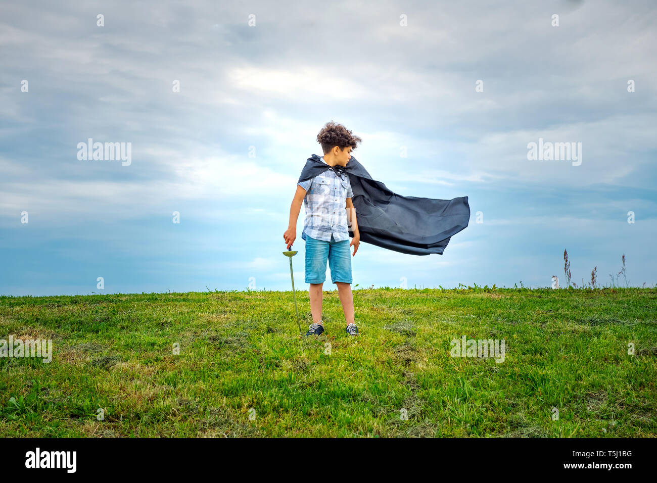 Ragazzo giovane super eroe con il mantello di soffiaggio in piedi all'aperto in un campo verde nel vento guardando dietro a guardarlo billow nel vento Foto Stock