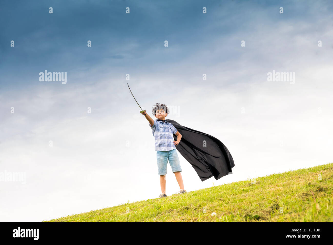 Orgoglioso ragazzo giocando il super eroe che indossa un mantello fatti in casa e brandendo una spada giocattolo sullo skyline contro un cielo blu Foto Stock
