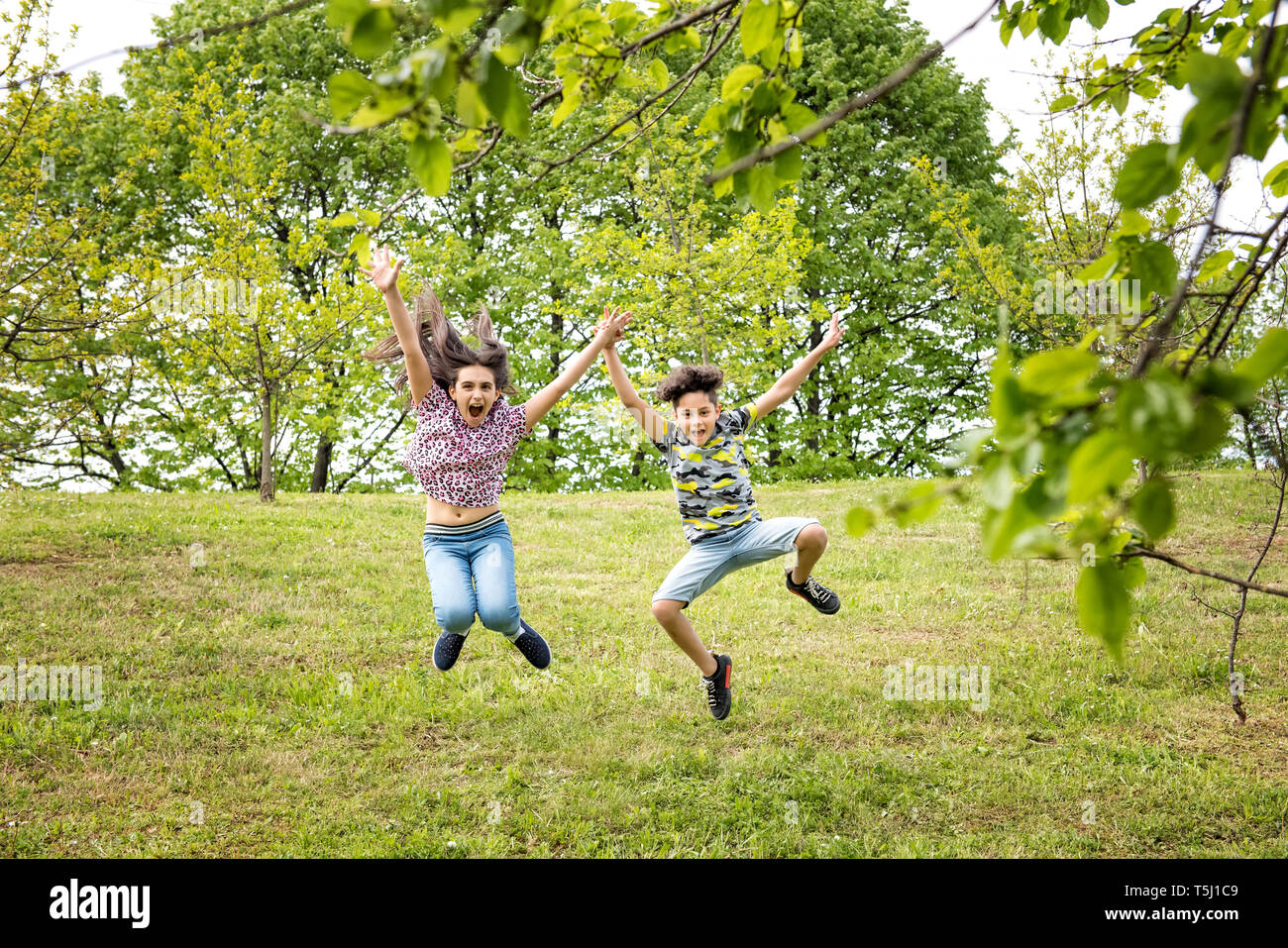 Carino energico giovane fratello e sorella saltando insieme in un parco alberato in primavera per celebrare le giornate più calde Foto Stock