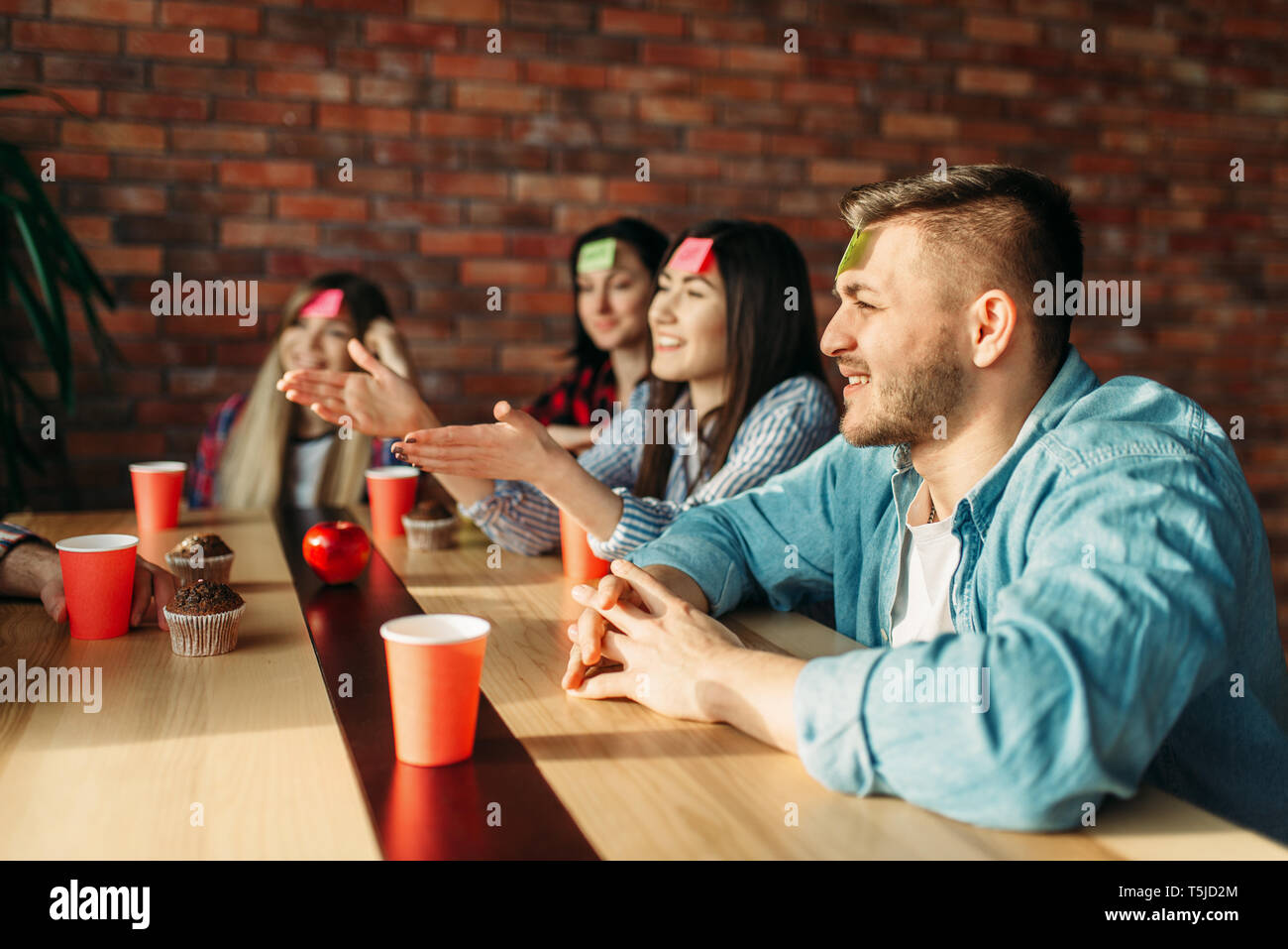 Sorridente amici giocando sticker note di fronte al gioco. Azienda svolge chi sono io a tavola in cafe, indovinare il personaggio Foto Stock