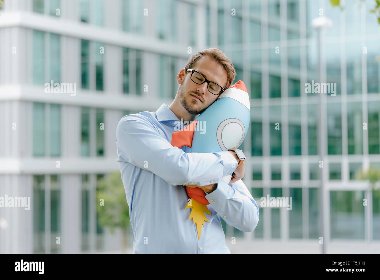 Giovane imprenditore in piedi di fronte a un ufficio moderno edificio, abbracciando razzo giocattolo Foto Stock