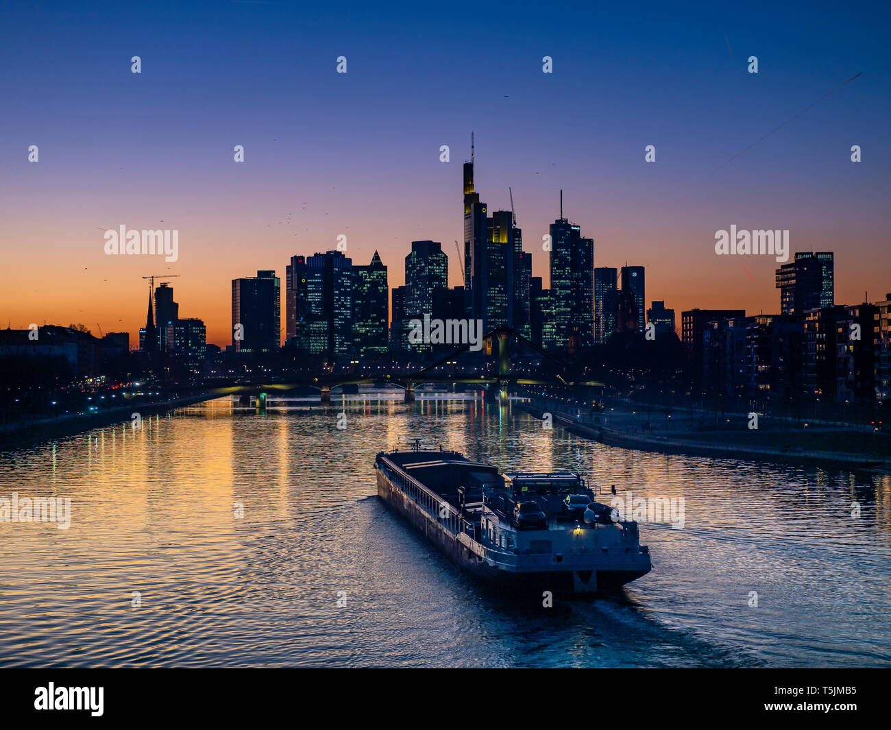 Germania, Hesse, Francoforte, Skyline del financial district, fiume principale e ponte Deutschherrn, nave da carico al tramonto Foto Stock