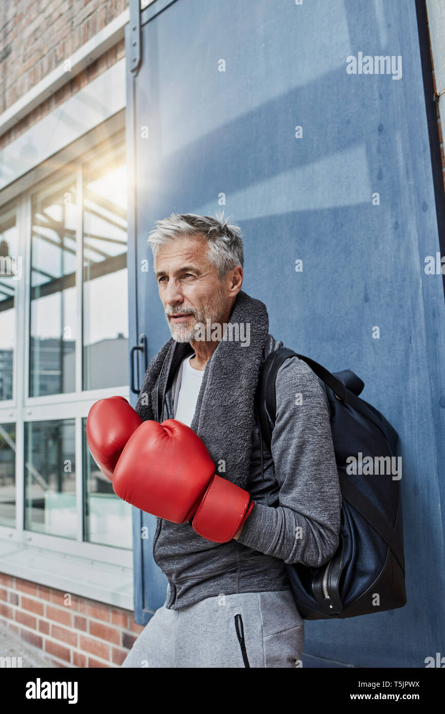Ritratto di uomo maturo con asciugamano, borsa sportiva e rosso guantoni da pugilato in piedi nella parte anteriore della palestra Foto Stock