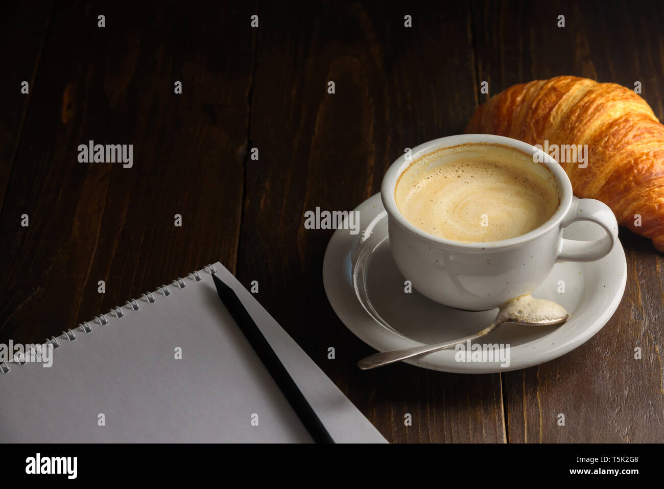 Cappuccino con croissant sul legno scuro dello sfondo. Spazio di testo. Vuoto il blocco note e matita vicino ad essa Foto Stock