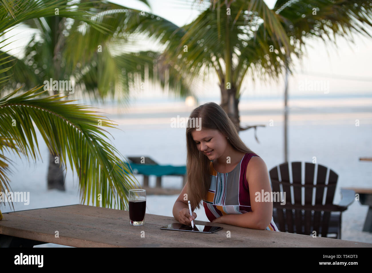 Giovane donna che indossa abiti utilizzando tablet sulla spiaggia al mattino sullo sfondo di palme in Florida con un bicchiere di soda Foto Stock Giovane donna che indossa abiti utilizzando tablet sulla spiaggia al mattino sullo sfondo di palme in Florida con un bicchiere di soda Foto Stock