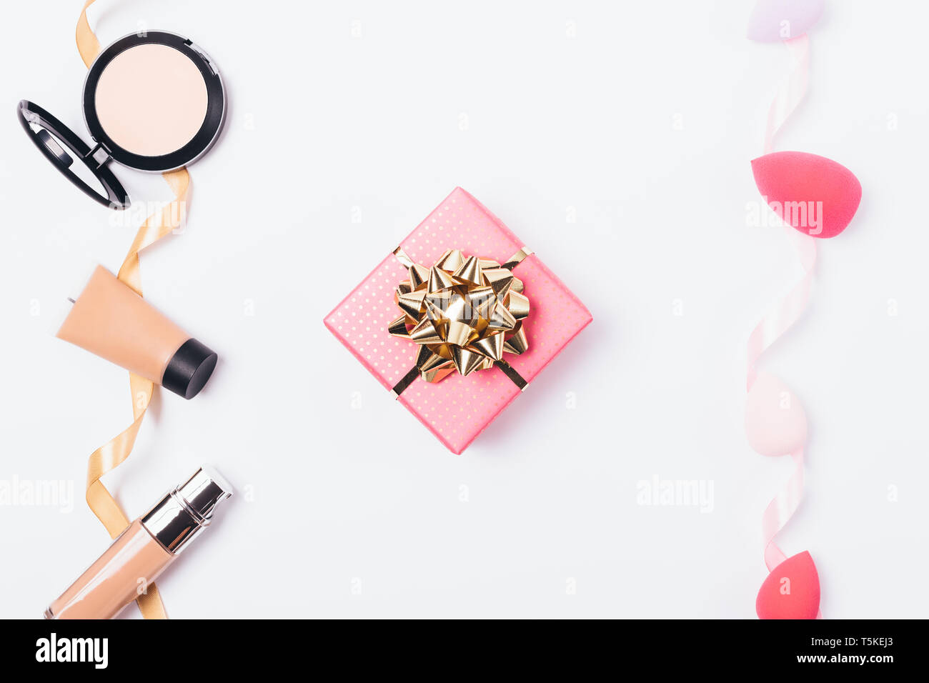 Rosa confezione regalo con golden bow tra cosmetica decorativa per affrontare anche la tonalità della pelle e il trucco spugne per la sua applicazione, vista dall'alto su sfondo bianco. Foto Stock