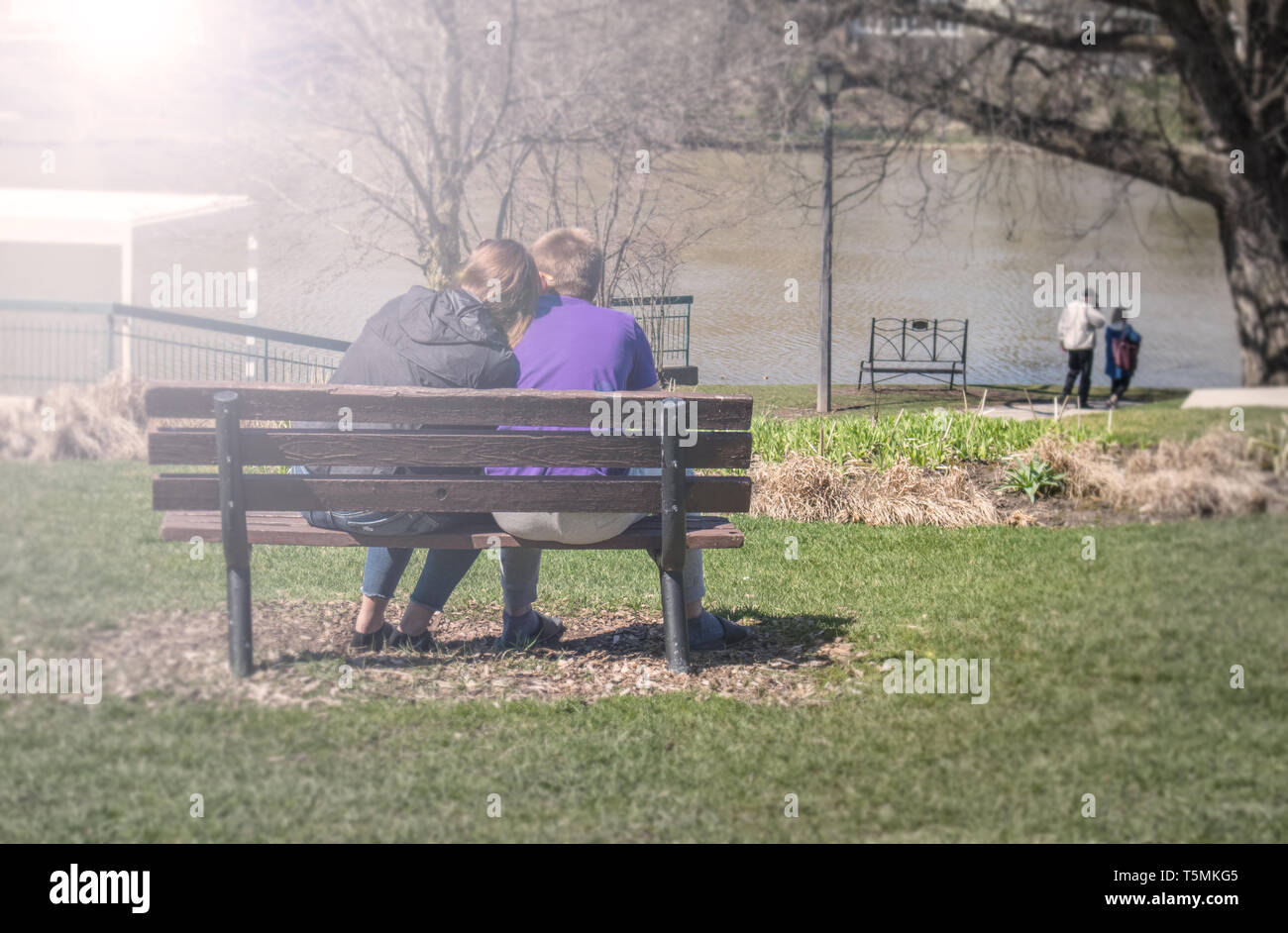 Un giovane seduto su una panchina nel parco. Ella teneramente stabilisce la sua testa sulla sua spalla, durante una giornata di sole di primavera. Vista da dietro. Foto Stock