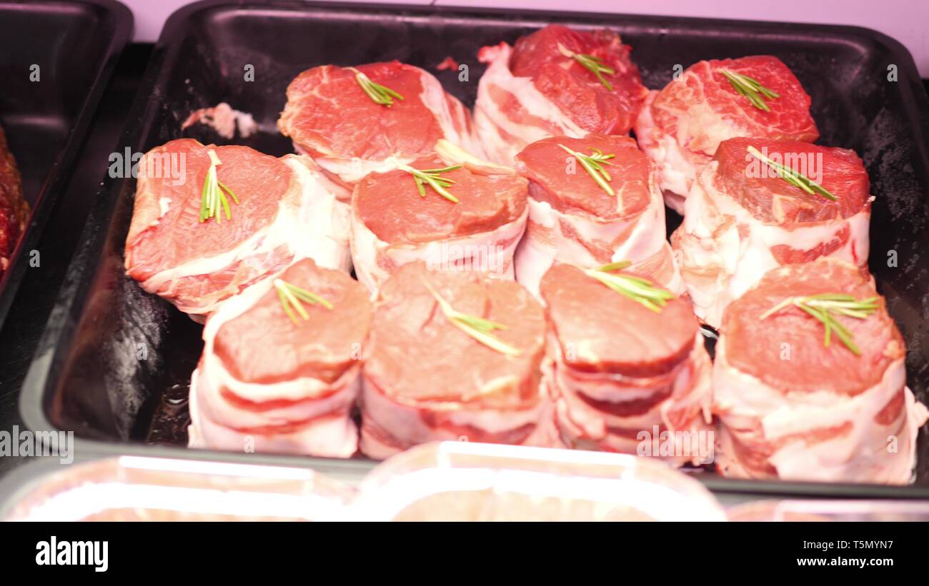 Pezzi di filetto crudo fresco di carne di manzo con rosmarino, carne di maiale e fette di bacon sono giacenti in vassoi di nero sullo scaffale del negozio pronti per la vendita in slow mo Foto Stock