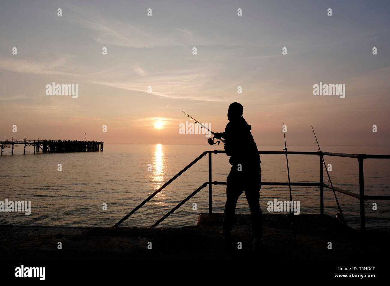 Pesca sportiva fisherman mediante aste di spiaggia espressi dal marciapiede al di sopra Totland Bay come il sole tramonta oltre il lontano orizzonte e Totland Pier. Foto Stock
