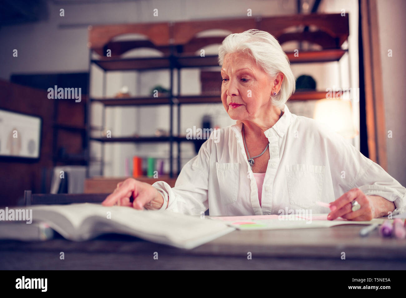 Elegante anziana signora con le rughe del viso seduta a tavola e libro di lettura Foto Stock