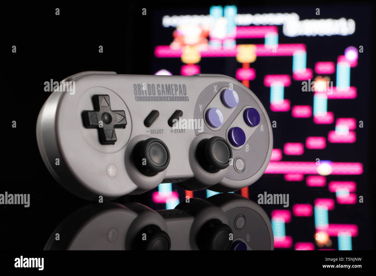 Un grigio, giochi wireless controller, seduto in posizione eretta su una superficie a specchio, di fronte a un fuori fuoco gioco arcade. Foto Stock