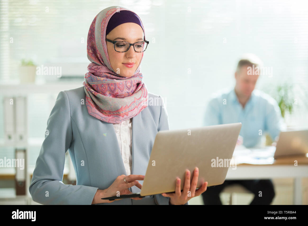 Bella donna elegante in hijab e occhiali in piedi in ufficio, tenendo il laptop. Ritratto di fiducioso imprenditrice musulmana. Ufficio moderno con grandi Foto Stock