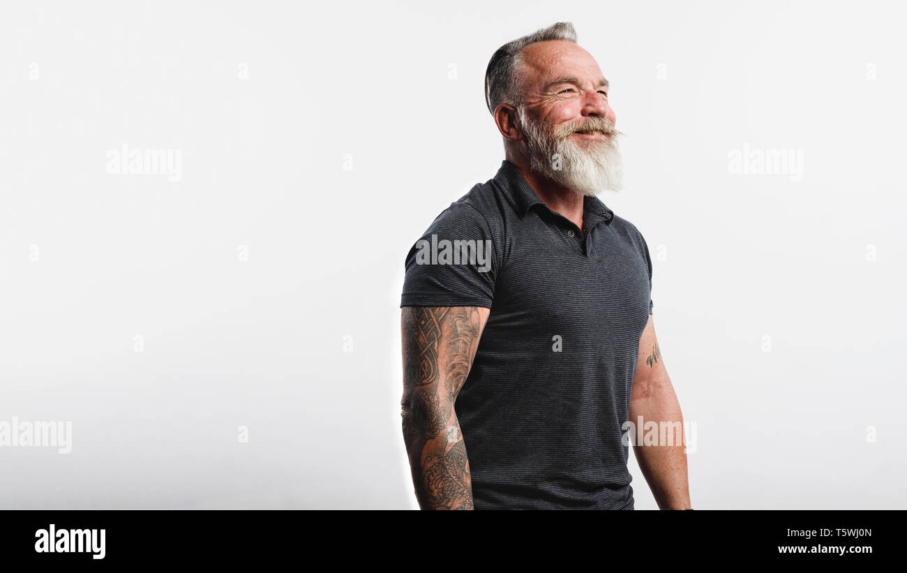 Felice l'uomo senior con tatuaggio su bracci in piedi contro uno sfondo bianco. Ritratto di vecchio maschio muscolare con la barba bianca che guarda lontano. Foto Stock