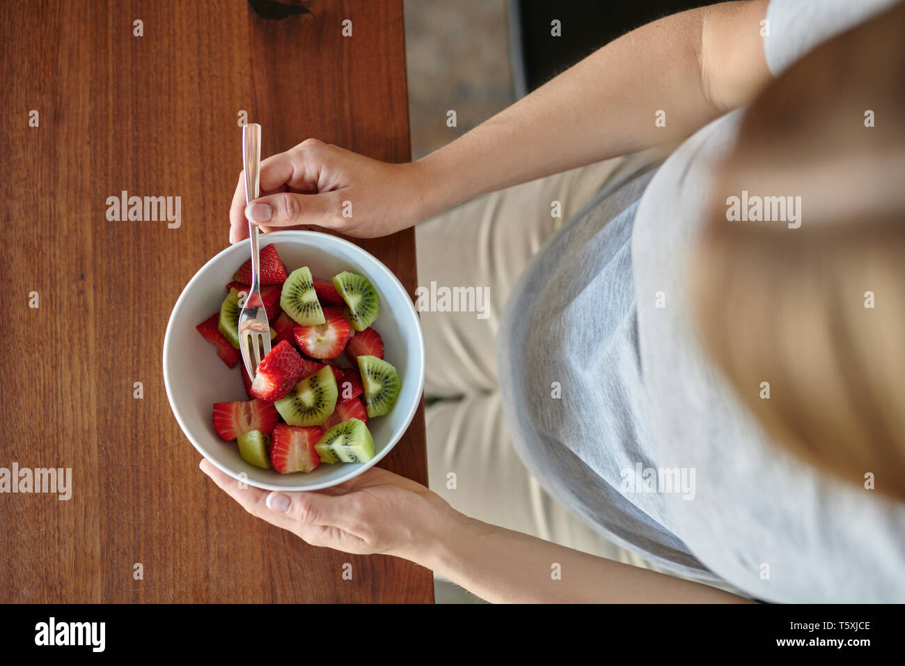 Donna incinta mangiare una tazza di macedonia di frutta con fragole fresche seduti a un tavolo da pranzo in un vicino sul cibo e la sua pancia in una dieta sana Foto Stock