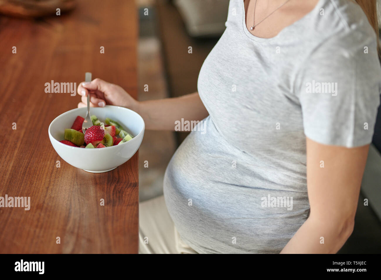 Donna incinta mangiare una tazza di macedonia di frutta con fragole fresche seduti a un tavolo da pranzo in un vicino sul cibo e la sua pancia in una dieta sana Foto Stock