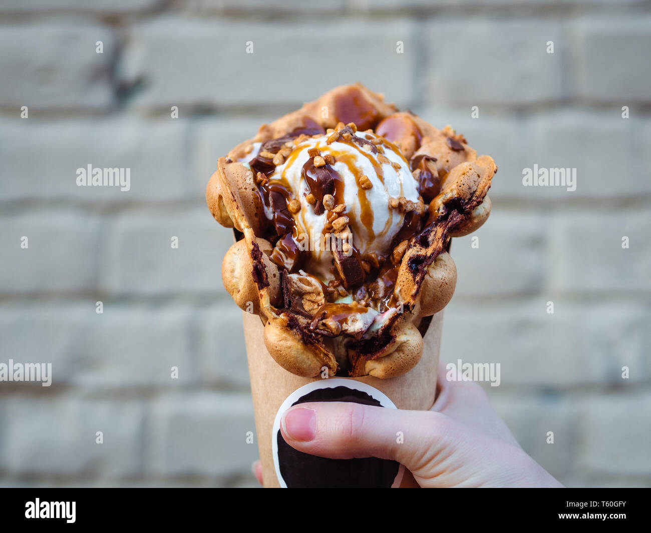 Hong Kong cialde in mano femmina sul muro di mattoni in background. Bolla di deliziosi waffle con gelato e sciroppo di caramello nella donna la mano. Cibo alla moda. Foto Stock