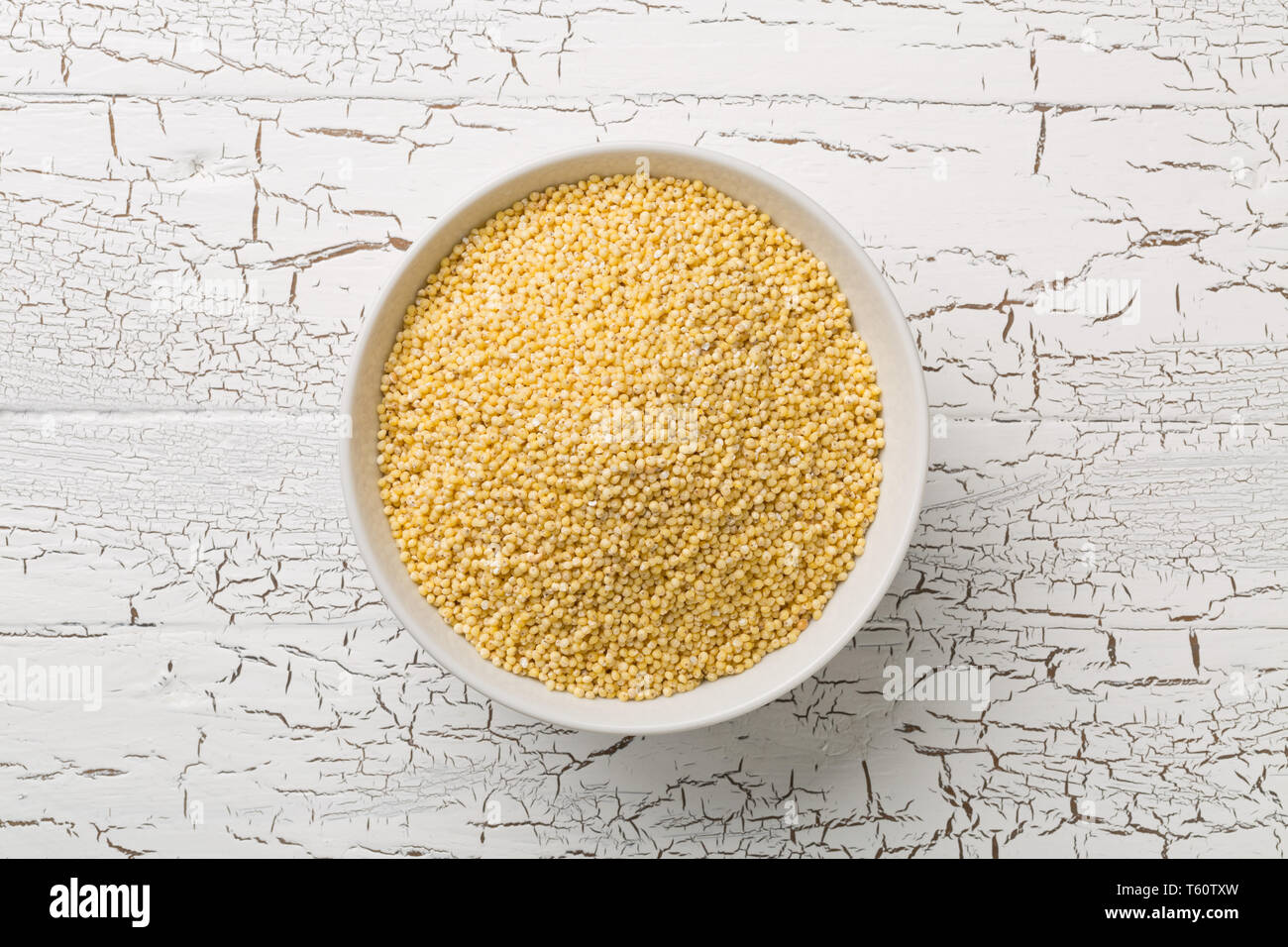 Pila di miglio dorato, un senza glutine di sementi di grano, nella ciotola bianco su bianco rustico sfondo tabella vista in pianta da sopra Foto Stock