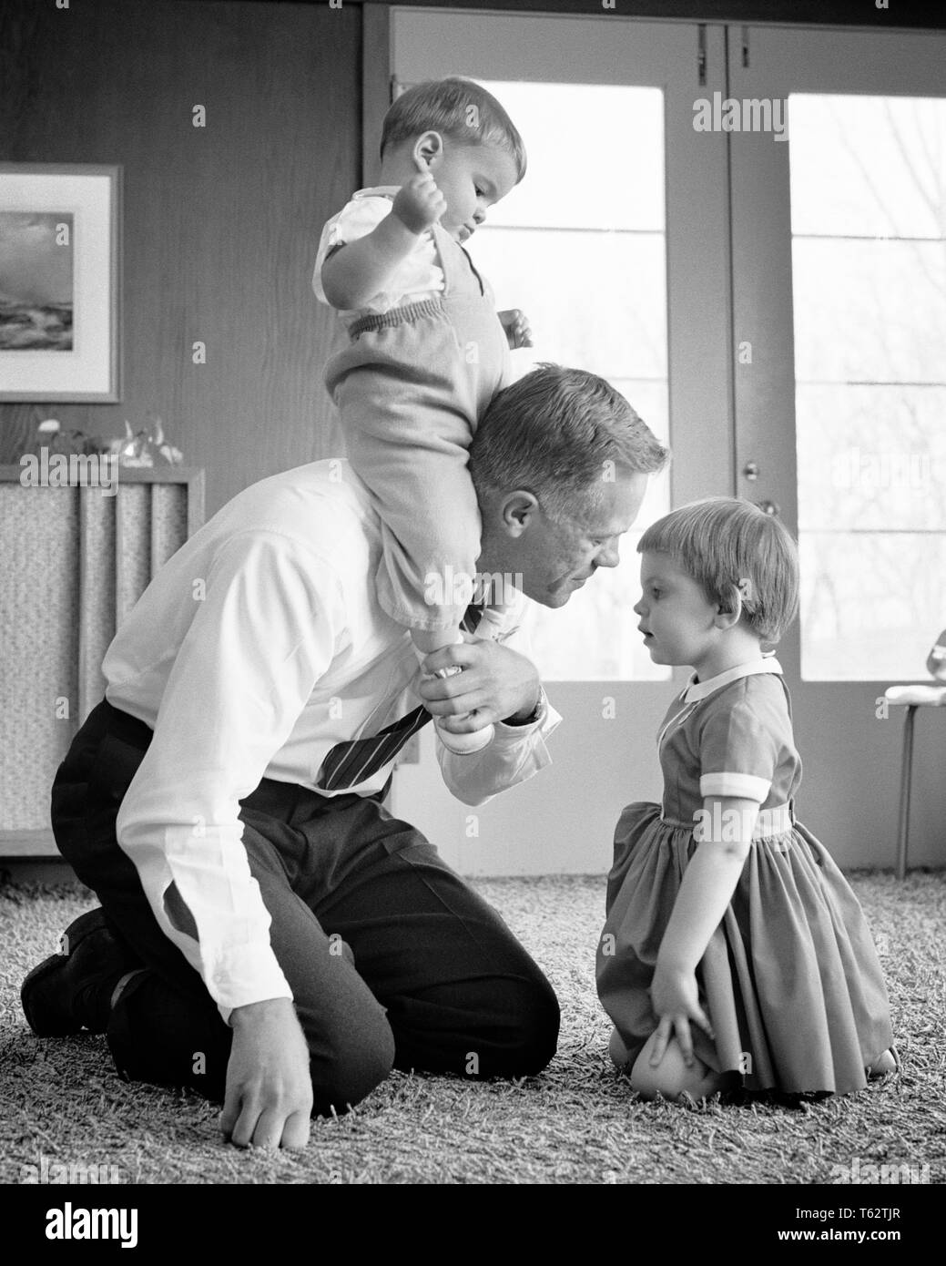 Anni sessanta padre giocando sul pavimento con bambini inginocchiato con TODDLER GIRL bambino seduto sulle sue spalle - J11792 HAR001 HARS SORELLA 1 bambini figli femmine PARENTING FRATELLI HOME VITA spazio copia amicizia a mezza lunghezza figlie persone cura maschi fratelli sorelle padri B&W in ginocchio la felicità del suo papà gemelli baby boy piggy back crescita novellame PIGGYBACK convivere in bianco e nero di etnia caucasica HAR001 in vecchio stile Foto Stock