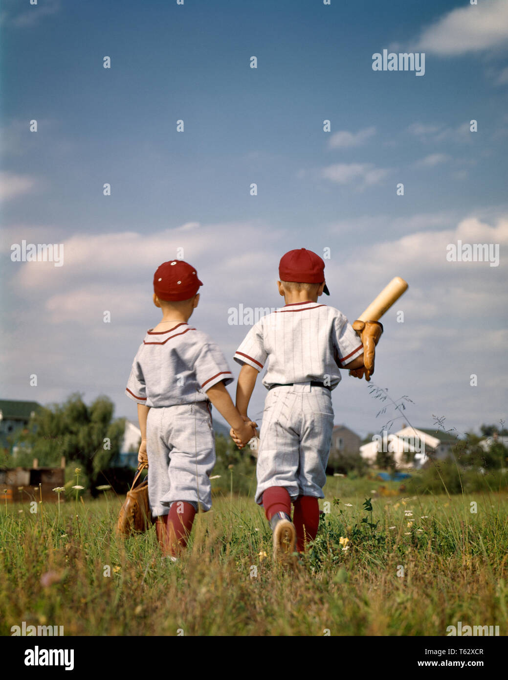 Anni sessanta due ragazzi fratelli indossando Little League Baseball uniformi tenendo le mani A PIEDI DALLA FOTOCAMERA - kj3545 HAR001 HARS FRATELLI spiritualità successo felicità Little League forza avventura VITTORIA MITT Holding Hands uniformi di pari livello concettuale di collegamento per andare a casa elegante supporto, lontano dalla telecamera di perdere il novellame affiatamento tappo a sfera fraterno etnia caucasica HAR001 in vecchio stile Foto Stock