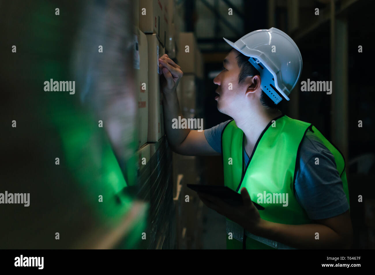 Vista laterale dei giovani asiatici magazzino uomo lavoratore con sicurezza elmetto è controllare i dettagli di un ordine con una tavoletta digitale alla camera di inventario Foto Stock