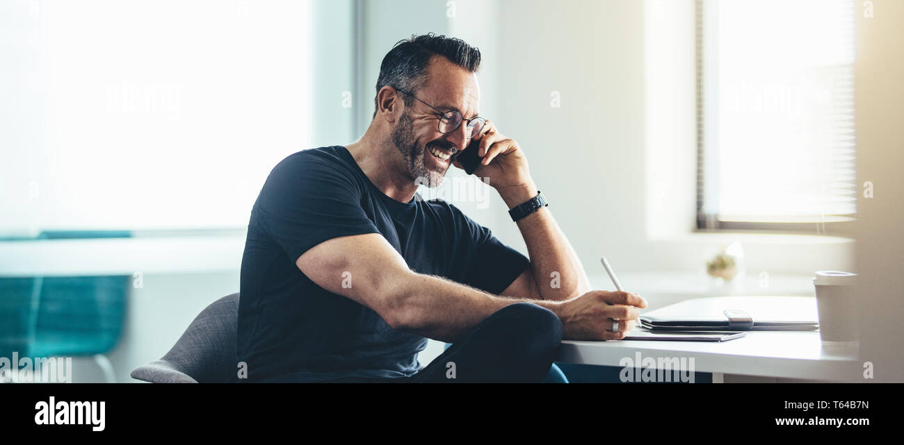 Uomo sorridente al lavoro parlando al cellulare e al lavoro sul suo digitale compressa. Allegro imprenditore lavora in ufficio. Foto Stock