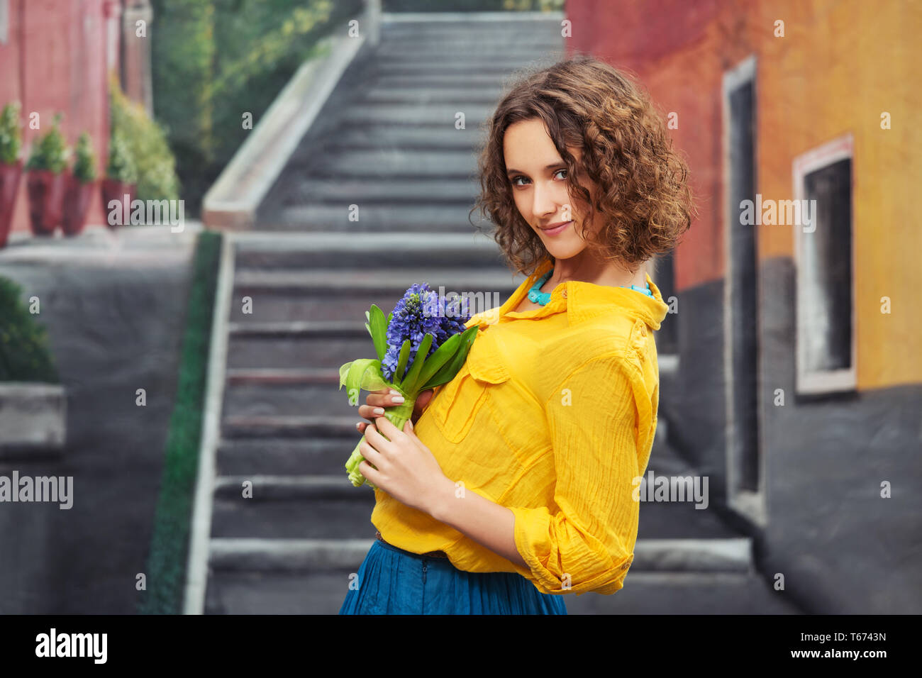 Felice moda giovane donna con bouquet di fiori a piedi nella città strada elegante modello femminile indossare magliette di colore giallo e la gonna blu Foto Stock