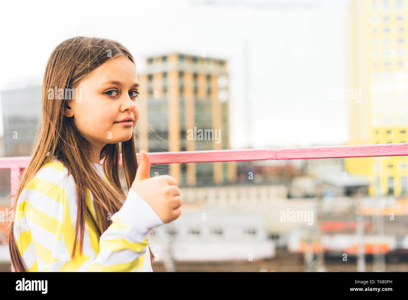 Una ragazza adolescente in un maglione giallo si erge contro lo sfondo del paesaggio urbano e mostra una mano con un dito sollevato Foto Stock