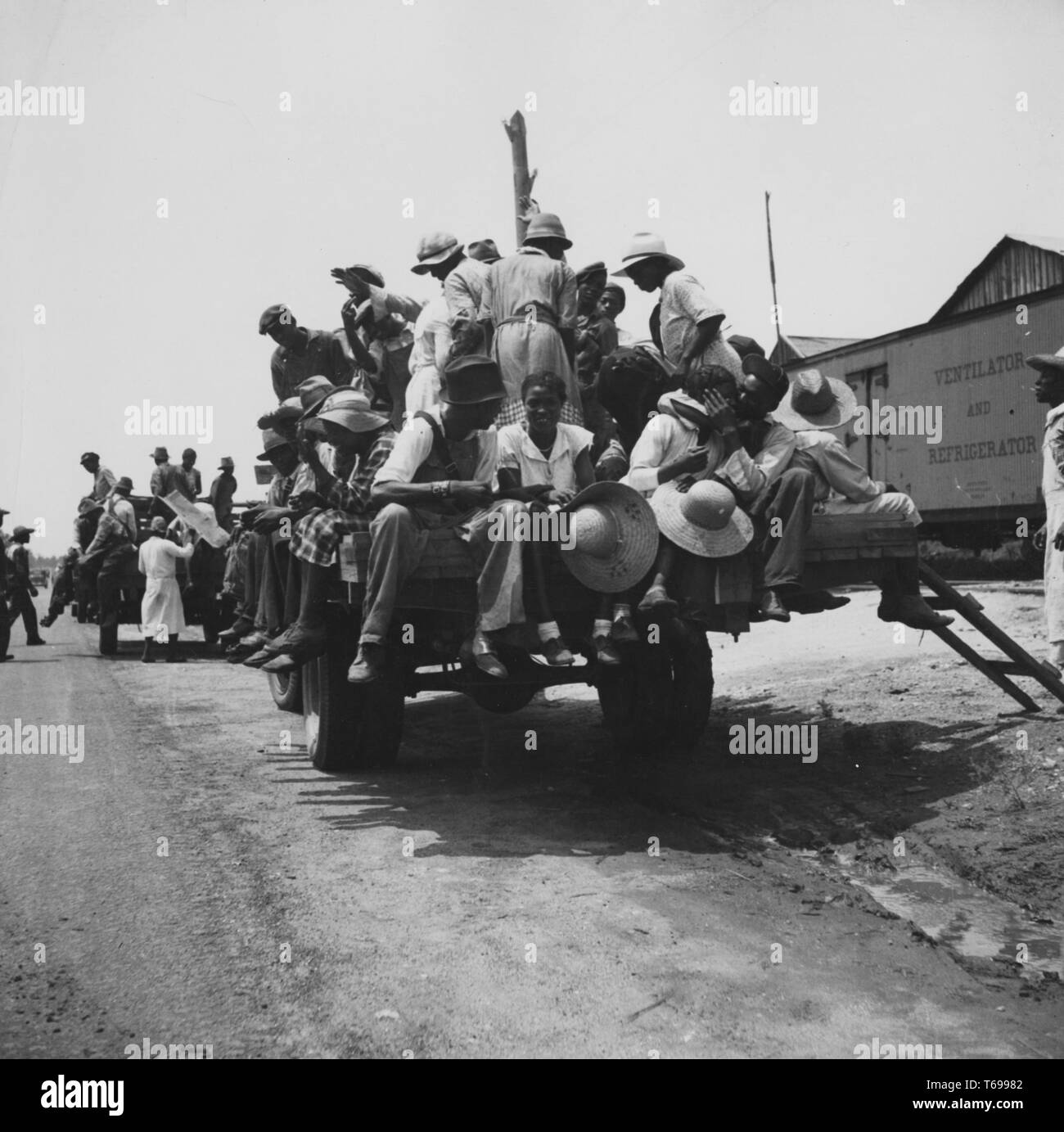 Fotografia in bianco e nero di afro-americano di uomini e donne di imbarco e seduta in carrelli dello scanner a superficie piana in rotta per i loro posti di lavoro come braccianti agricoli su una piantagione di pesche situato in Muscella, Georgia, USA, con una vista in primo piano di quattro persone sedute sulla parte posteriore del carrello, compreso un uomo che indossa un piccolo colmato hat, una donna sorridente tenendo un grande cappello di paglia e un uomo che abbraccia una donna; fotografato da Dorthea Lange sotto il patrocinio degli Stati Uniti " Farm Security Administration, 1936. Dalla Biblioteca Pubblica di New York. () Foto Stock