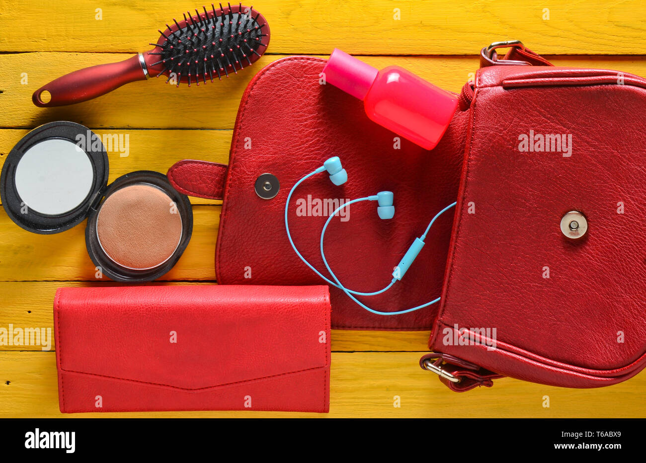 Qual è la donna della borsa? Un sacchetto rosso, portafoglio in pelle, cuffie, specchio, pettine, bottiglia di profumo su un giallo tavolo in legno. Vista dall'alto. Foto Stock