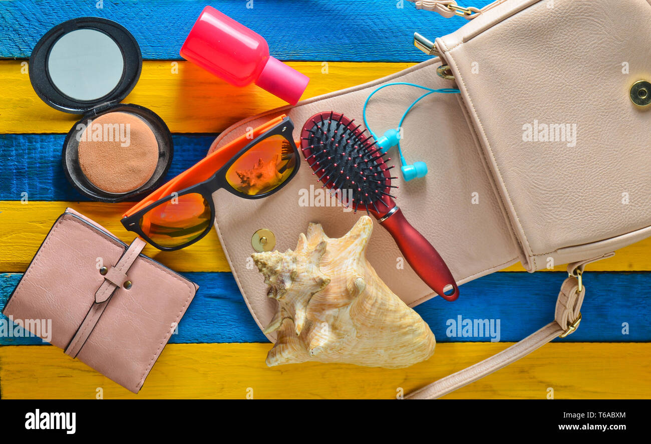 Qual è la donna della borsa? Estate spiaggia accessori, shell. Un sacchetto, portafoglio in pelle, cuffie, specchio, pettine, bottiglia di profumo su un giallo blu sulla scheda di legno Foto Stock