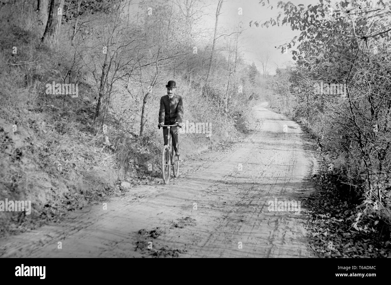 Un Bowler cappello uomo corse la sua bici giù per una strada di campagna in Pennsylvania, ca. 1900. Foto Stock