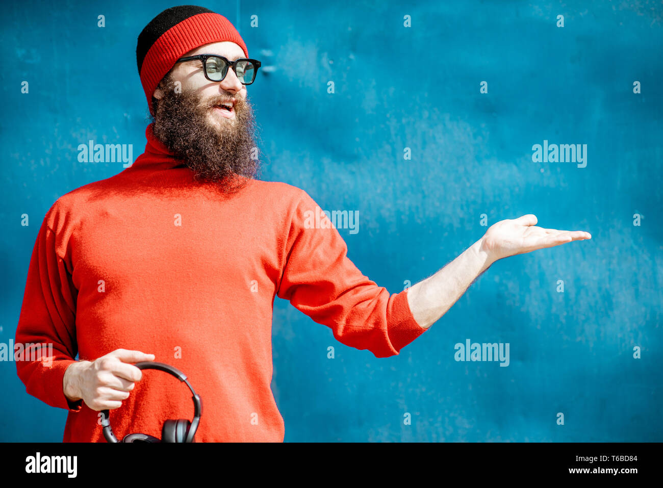 Ritratto di un elegante uomo barbuto vestito di rosso maglione e hat mostra su sfondo blu Foto Stock