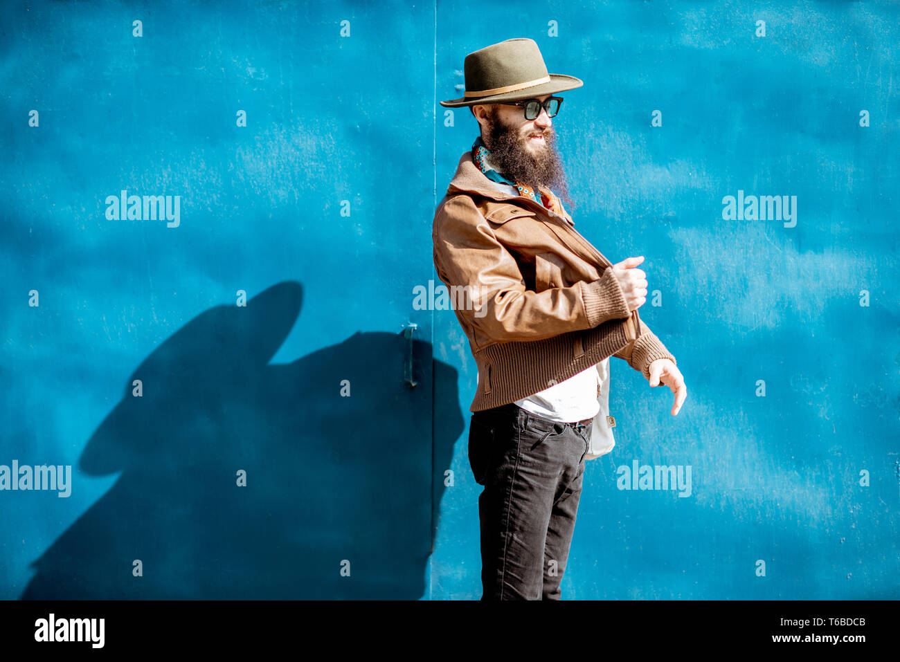 Ritratto di un elegante uomo barbuto vestito in giacca e hat in piedi con la borsa su sfondo blu all'aperto Foto Stock