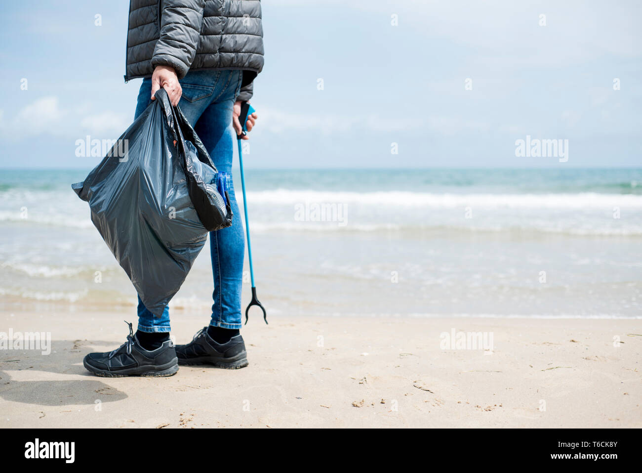 Primo piano di un uomo caucasico raccogliere rifiuti con un cestino con terminazione a molletta per stick, su una solitaria spiaggia, come un intervento per pulire l'ambiente naturale, con s Foto Stock