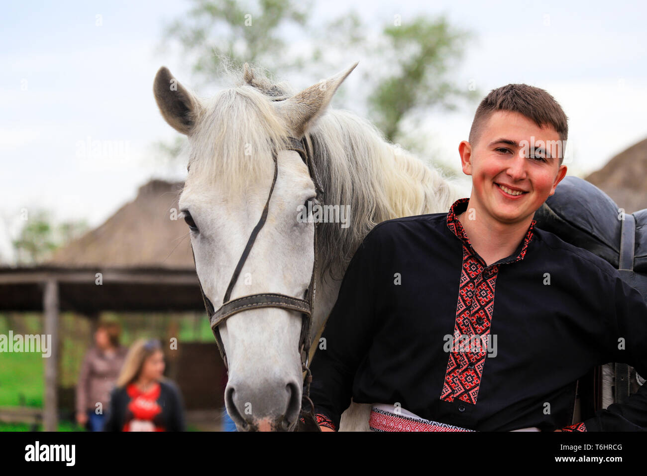 Petrikovka, Dnepropetrovsk regione, Ucraina, 28 04 2019 un giovane cosacco ucraino in camicia ricamata sorrisi e trattiene un cavallo grigio. Equitazione Foto Stock