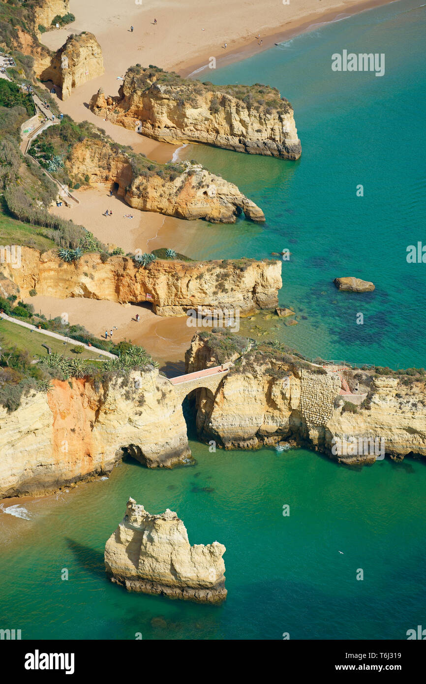 VISTA AEREA. Serie di scogliere parallele sul mare a Praia Dos Estudiantes (spiaggia degli studenti), ponte romano in primo piano. Lagos, Algarve, Portogallo. Foto Stock